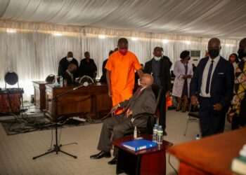 Ex-president Guebuza, seated, talks to his son Ndambi Guebuza, wearing an orange prison uniform. The younger Guebuza is a defendant in the long-awaited trial, which is unfolding in a marquee in a high-security jail | AFP